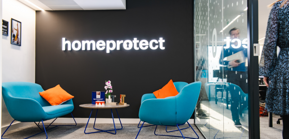 A modern office, with two blue chairs around a small table, with orange pillows. The black wall has "Homeprotect" written in a white neon sign